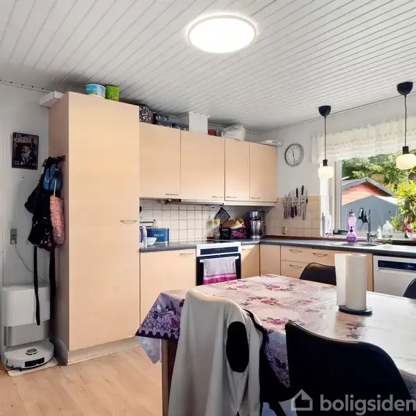 Køkken med lyse trækøkkenskabe, komfur og vask ved hjørnevindue. Bord med dug i midten, omgivet af stole. Vinduet viser en have udenfor. Ingen tekst fundet.