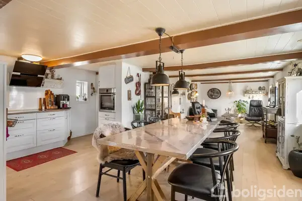 Langt spisebord med stålben står i åbent køkken-alrum, omgivet af sorte stole. Køkkenskabe til venstre, hvide lofter med træbjælker og opholdsområde i baggrunden. Lyse og indbydende omgivelser.