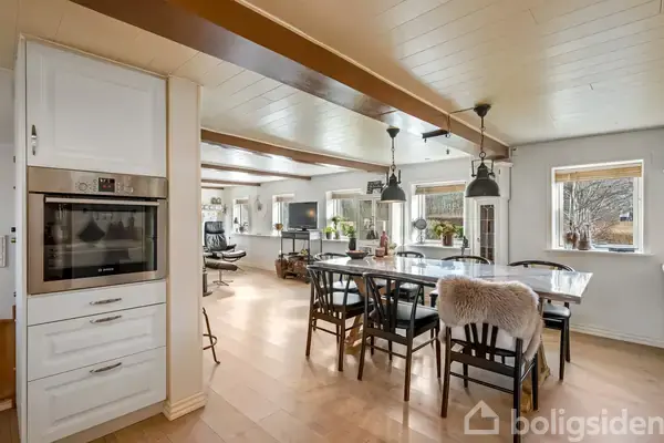 Ovn indbygget i et hvidt skab, omgivet af en åben køkken-stue, hvor der står et spisebord med stole under hængende lamper. Vinduer bader rummet i naturligt lys.