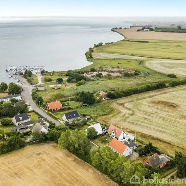 En klynge af huse ligger roligt ved en bugt med en lille bådehavn. Omgivet af marker og grønne områder skaber det en idyllisk, landlig atmosfære nær vandet.