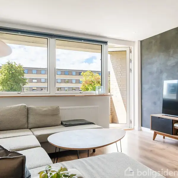 En grå hjørnesofa står i en lys stue med store vinduer. Et rundt sofabord er foran, og et fladskærms-tv er placeret på et tv-møbel.