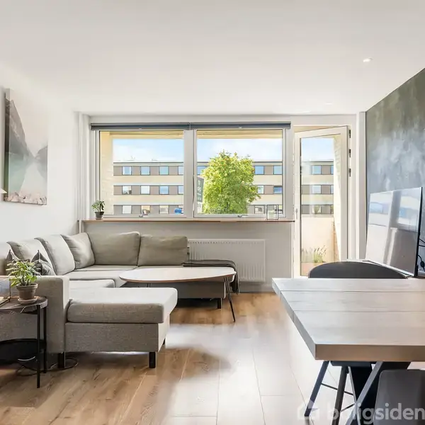 En lys stue med en grå hjørnesofa ved et vindue. Til højre står et spisebord, blomster i en vase, og et tv op ad en mørk væg.