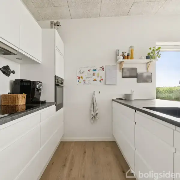 Hvidt køkken med sorte bordplader, hvor en kaffemaskine står ved siden af en kurv. Væggen har opslagstavler og hylder. Stort vindue med udsigt over grønt landskab.