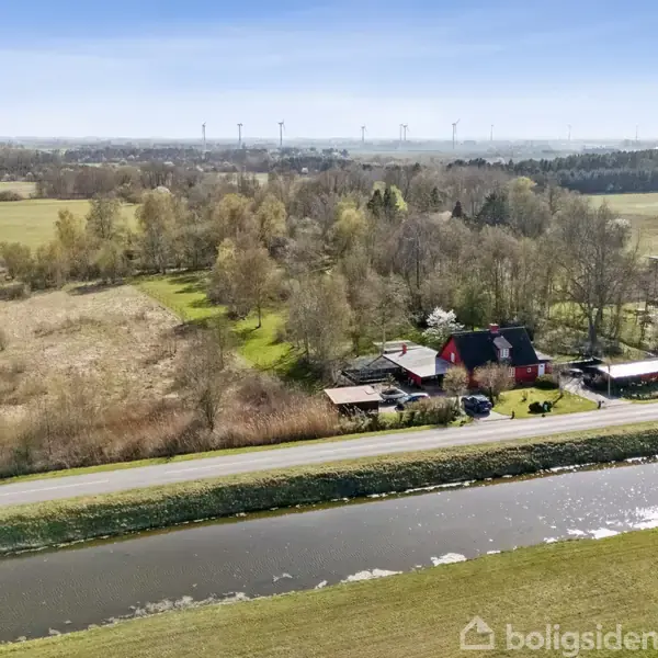 Landhus med sort tag beliggende ved en landevej. Omgivet af træer og grønne marker, med en kanal i forgrunden. Vindmøller kan ses i det fjerne.