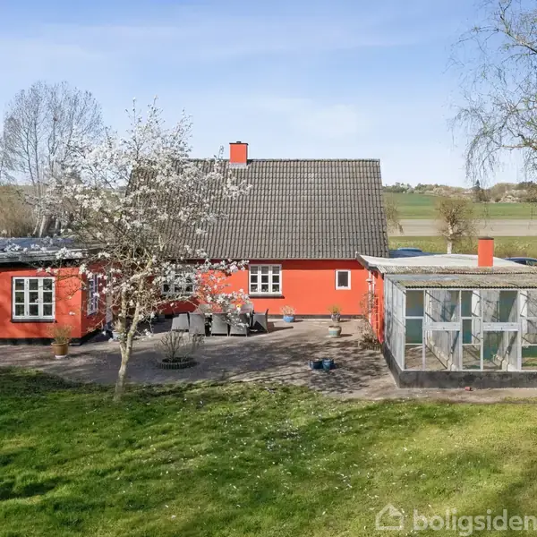 En rødmuret hus står omgivet af en stor have med blomstrende træer. Ved siden af huset er et drivhus. Landskabet i baggrunden viser åbne marker og skovområder.