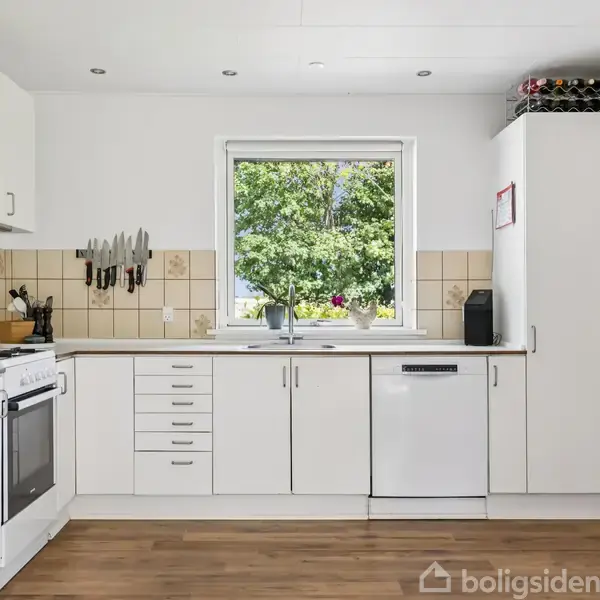 Et hvidt køkken med komfur og opvaskemaskine langs væggen. Knive hænger over køkkenbordet. Udsigt til en grøn have gennem vinduet. Brune fliser danner baggrund.