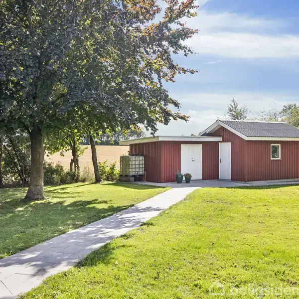 Et rødt skur med et gråt tag står blandt grønne træer og en velholdt græsplæne. En flisebelagt sti fører til skuret, der ligger under en klar blå himmel.