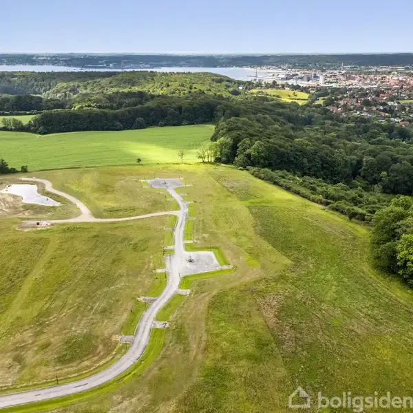 Grønne marker med en snoet vej fører til en skovkant. I baggrunden ses en by ved en kystlinje med blå himmel over.
