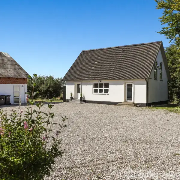 Hus med skråt tag står på en grusbelagt gårdsplads, omgivet af grønne buske og træer under en klar blå himmel. En stor garagebygning ses til venstre.