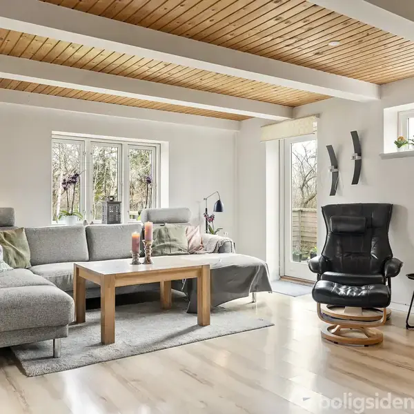 Grå sofa med puder ved stuebord med stearinlys, placeret i et lyst lokale med trægulv og træloft. Store vinduer giver udsigt til have, og en sort lænestol står ved siden.