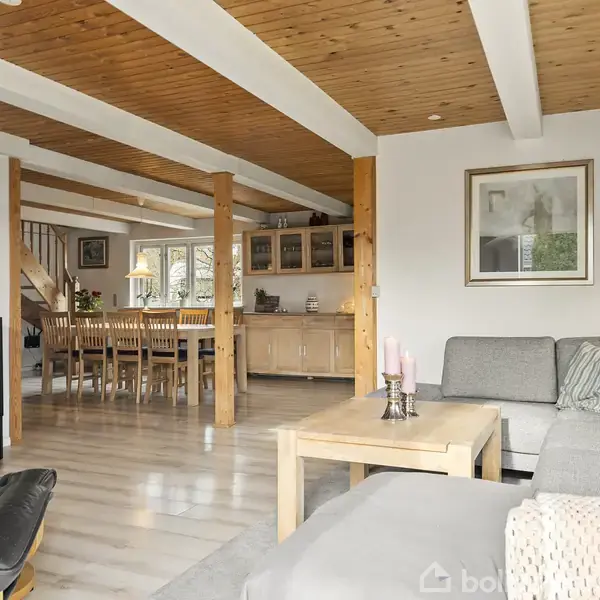 Grå sofa står i stue med bord, lyse trævægge og loftsbjælker. Et spisebord med stole ses i baggrunden nær trappe. Væggene har billeder, og et vindue giver lys.
