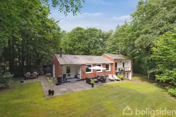 Et rødt murstenshus ligger omgivet af frodig skov. Foran huset er der en græsplæne med en terrasse, der har udendørs møbler og parasoller.
