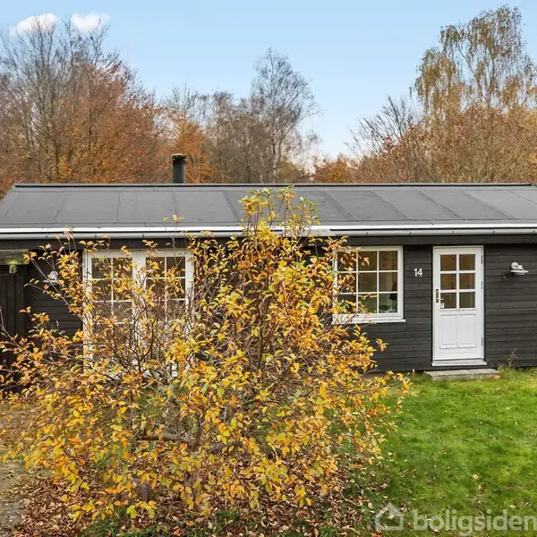 Et sort træhus med hvide vinduer og dør, mærket "14," står foran et farverige efterårstræer. En gårdsplads med brosten er delvist synlig foran huset.
