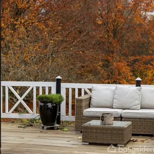 En udendørs sofa med hvide hynder står på en træterrasse. Ved siden af er et lille bord og en plante i en sort potte. Baggrunden viser efterårsfarvede træer.