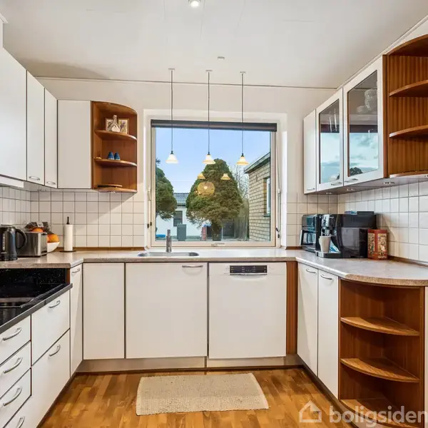 Hvidt køkken med trægulv; køkkenapparater inkluderer ovn, opvaskemaskine og mikroovn. Stor vindue med udsigt over grønne træer, omgivet af hvide skabe og træhylder. Pendellamper hænger ned.