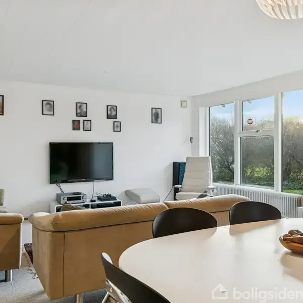 En stue med en beige hjørnesofa og tv på væggen. Et spisebord med stole står i forgrunden. Store vinduer lukker naturligt lys ind, omgivet af vægge med dekorationer.