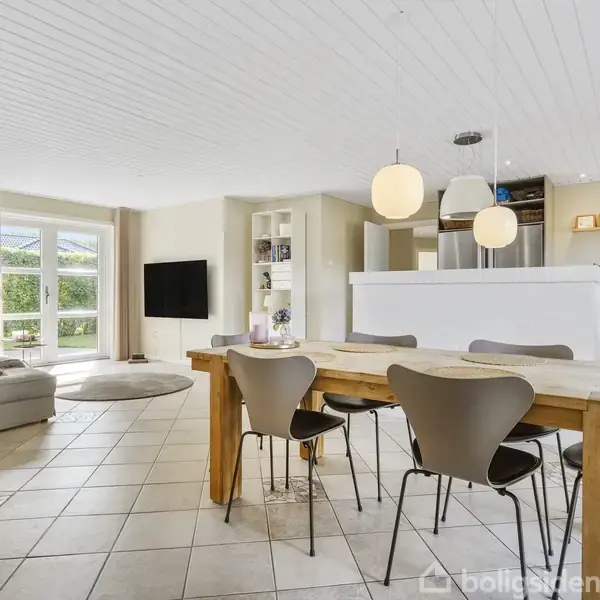 Et lyst, åbent køkken-alrum med spisebord og stole i forgrunden, sofa og TV ved vinduer i baggrunden. Indretningen er moderne med hvide lofter og flisegulv.