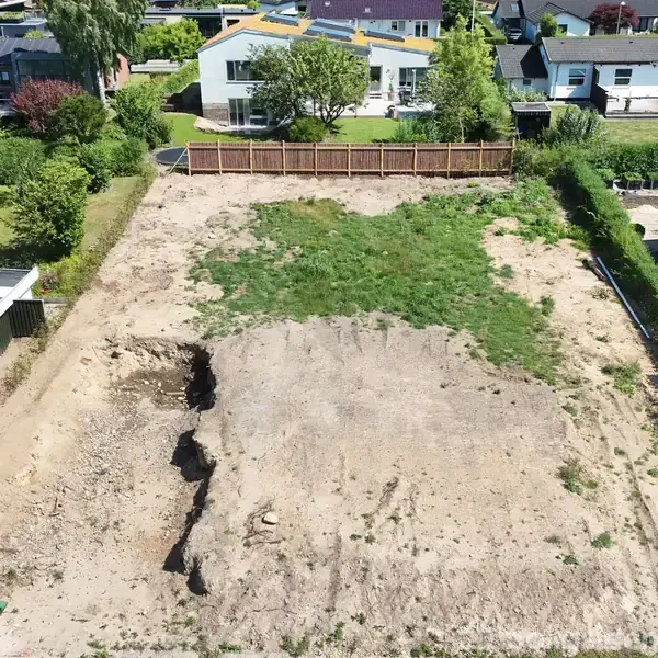 Byggegrund uden bygning, delvist overgroet med græs, omgivet af hække og huse i et boligkvarter. Fladt terræn med jord og sparsomme planter i en beboelsesvilla omgivelse.