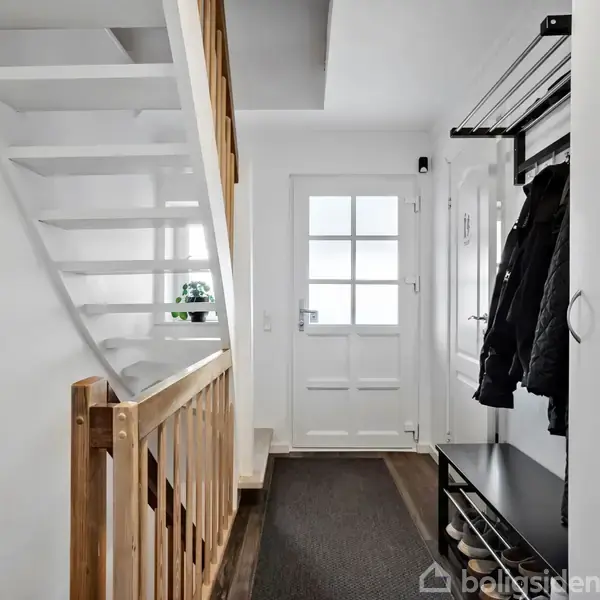 Trappe leder opad med hvidmalet gelænder. Entré i minimalistisk stil med stumtjener og bænke op ad væggen. En dør med glasruder fører ud, planter dekorerer vindueskarme.