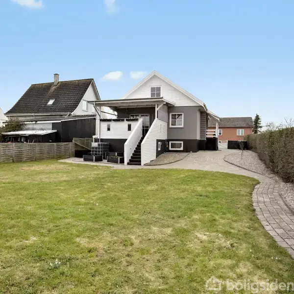 Et hvidt hus med trappe til veranda. Foran er en stor græsplæne og en flisebelagt sti langs hækken. Naboer ses i baggrunden under en blå himmel.