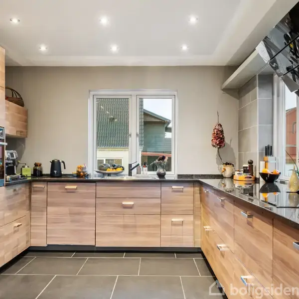 Moderne køkken med træskabe og sorte bordplader. Diverse køkkenapparater står på bordet, mens et stort vindue giver udsigt til udendørs bygninger. Flisegulvet er mørkt.