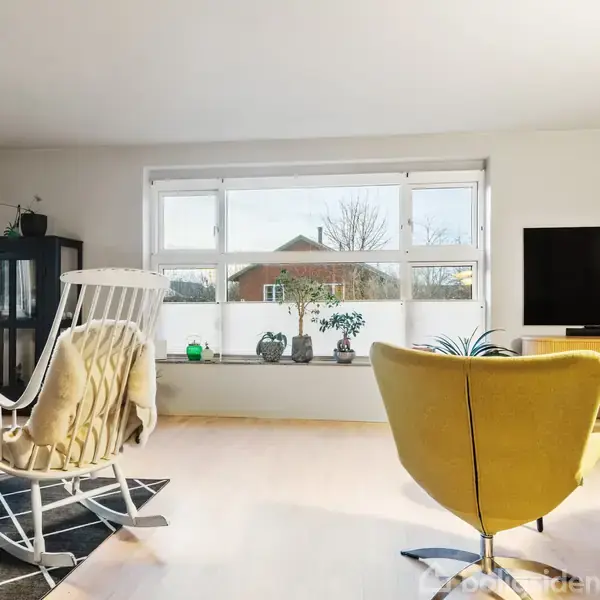 En stue med en gyngestol med tæppe, en gul lænestol og et tv. Et stort vindue bagved viser udsigt til en bygning. Stearinlys og planter dekorerer rummet.