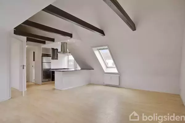 Et køkken-alrum med skrå loftsbjælker og stort ovenlysvindue. Køkkenet har en sort bordplade og skinnende emhætte. Rummet er lyst med lyse trægulve og hvide vægge.