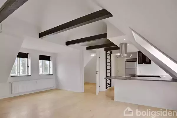 En lys stue med hvide vægge og træbjælker, store vinduer og et moderne køkken med indbygget ovn og emhætte. Trægulv og skrålofter skaber en rummelig atmosfære.