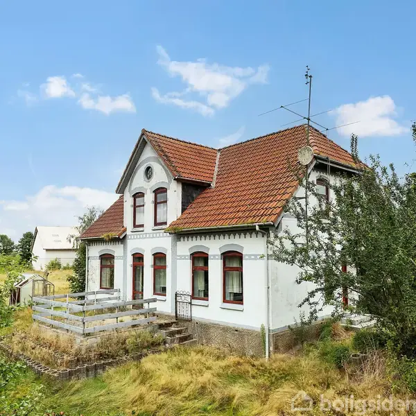 Et charmerende hvidt hus med rødt tegltag står i en frodig have med træer og buske. Huset har flere vinduer og en lille terrasse foran. Himlen er blå med få skyer.