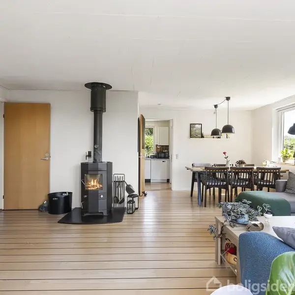 Brændeovn opvarmer stuen, hvor en sofa og spisebord er placeret. Store vinduer og lyse trægulve giver lys til rummet. Fjernsynet står i hjørnet.