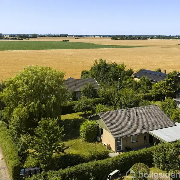 En lille, beige villa ligger roligt omgivet af grønne buske og træer. I baggrunden strækker gyldne marker sig ud under en blå himmel. En rød bil holder på vejen.