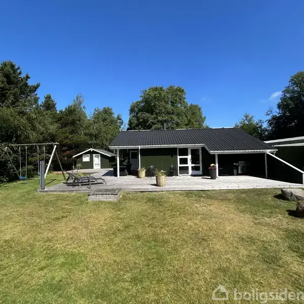 Et sort træhus står på en solrig græsplæne med en terrassedæk foran. En gyngestativ er til venstre, og grønne træer omkranser området.