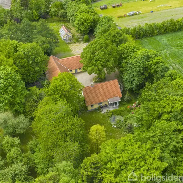 Et hus med rødt tag ligger omgivet af tæt skov og grønne marker. Et lille udhus er synligt i baggrunden.
