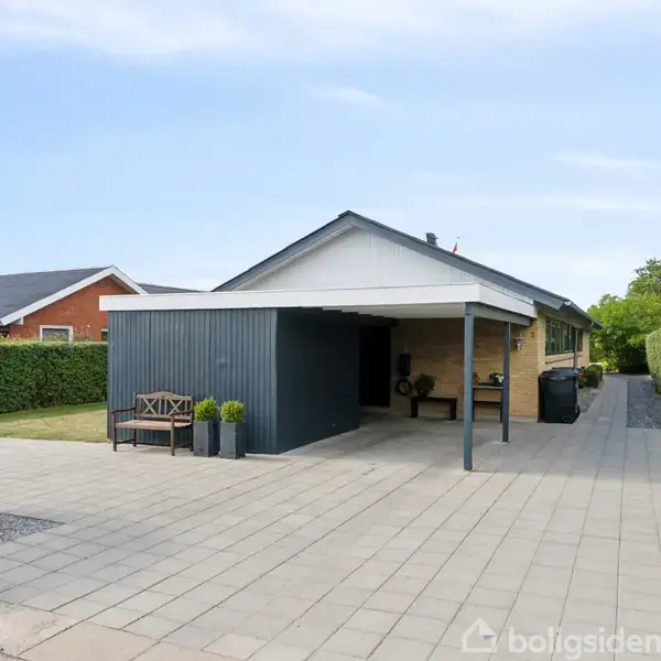 Et hus med carport står stille i en velholdt indkørsel omgivet af hække og græsplæne. En bænk og planter pryder det lille område foran carporten.