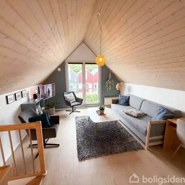 Loftstue med skrå træloft, en grå sofa, skrivebord og stol, rektangulært sofabord, tæppe og fjernsyn. Store vinduer med udsigt til røde huse udenfor.