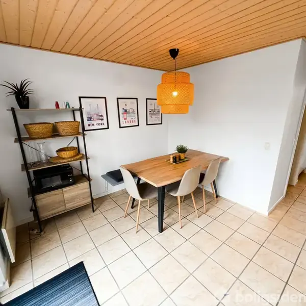 Et spisebord med fire stole står i et køkken med flisegulv. En orange lampe hænger over bordet. Hylde til venstre indeholder kurve og dekorationer. På væggen hænger billeder.