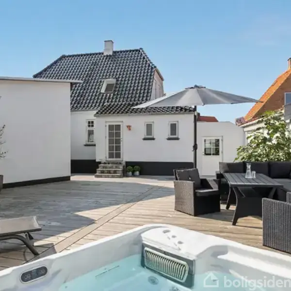 En udendørs terrasse har en jacuzzi i forgrunden. Ved siden af står et havesæt med parasol. Baggrunden viser en bygning med sort tag i en indhegnet gård.