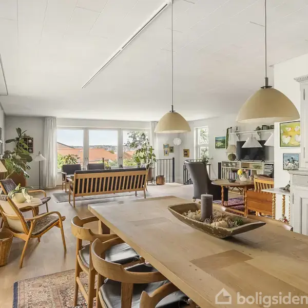 Spisestue med træbord og stole, omgivet af lys og hygge fra store vinduer og lamper. Baggrunden viser en stue med sofa, reoler og planter, skaber en indbydende atmosfære.
