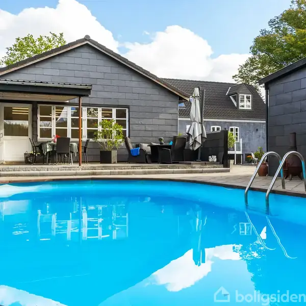 En blå swimmingpool står stille, omkranset af fliser foran et moderne, gråt hus med terrassemøbler og planter. Himlen er let overskyet, mens grønne træer rammer scenen ind.