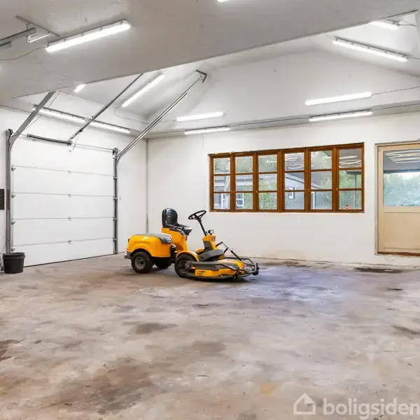 En gul plæneklipper står parkeret i en rummelig garage. Garagen har hvide vægge, store vinduer og en åben garageport. Der er også en sort bilfælg og et elektrisk kabel.