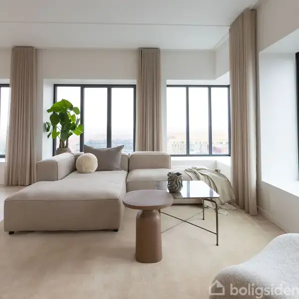 En beige sofa står i en lys stue med store vinduer og grå gardiner. En grøn plante og et rundt bord ses ved siden af sofaen på tæppet.
