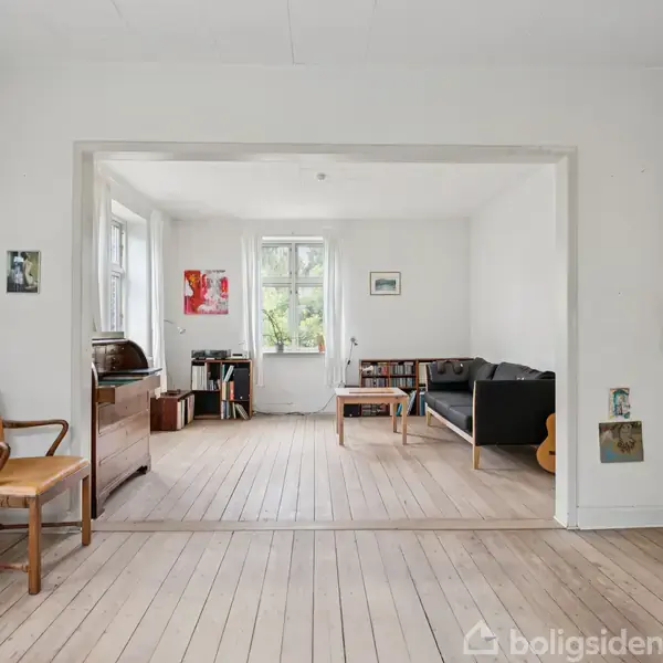 Lyse trægulve i en rummelig stue, hvor en sort sofa står mod højre væg. Der er et brunt bord ved siden af vinduet, og en lys træstol ved et bord til venstre.