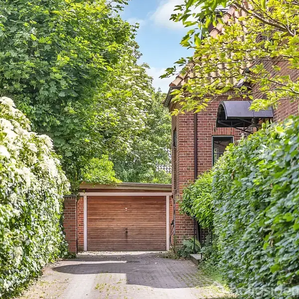 Garage med brun dør står lukket, omgivet af tætklippede, grønne hække og træer, ved siden af en brun murstensbygning i solbeskinnet have.