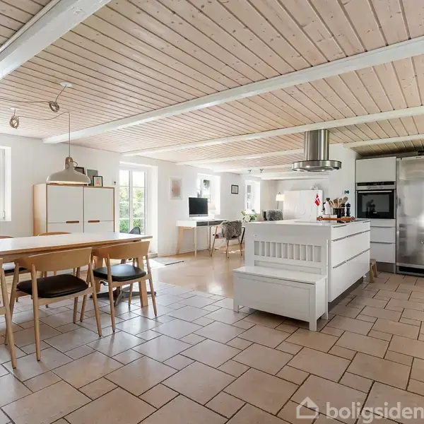 Et rummeligt køkken med spisebord og stole. Et hvidt køkkenbord står i midten, flankeret af køkkenmaskiner. Store vinduer og træloft skaber en lys atmosfære.