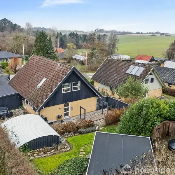 Et sort-hus står roligt omkranset af en velplejet have. Det ligger i et landligt område med flere omkringliggende huse og udsigt til åbne marker i baggrunden.