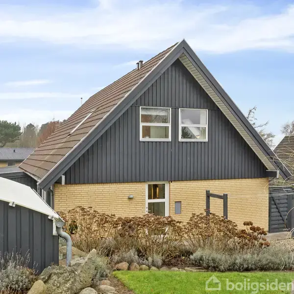 Et hus med mørkegrå gavl og røde tagsten står i en have. Græsplænen er grøn, omkranset af sten og planter, med enkelte træer i baggrunden under en blå himmel.