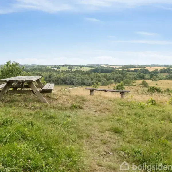Et træ-picnicbord står på en græsplæne med en bakket, grøn landskab i baggrunden. Solrigt vejr og blå himmel skaber en fredfyldt atmosfære.