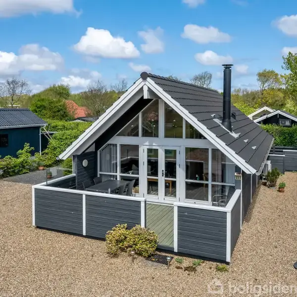 Et gråt sommerhus står stille omgivet af en grusbelagt gårdsplads og grønt buskads. Husets forside har store vinduer, og en terrasse omkranser bygningen.
