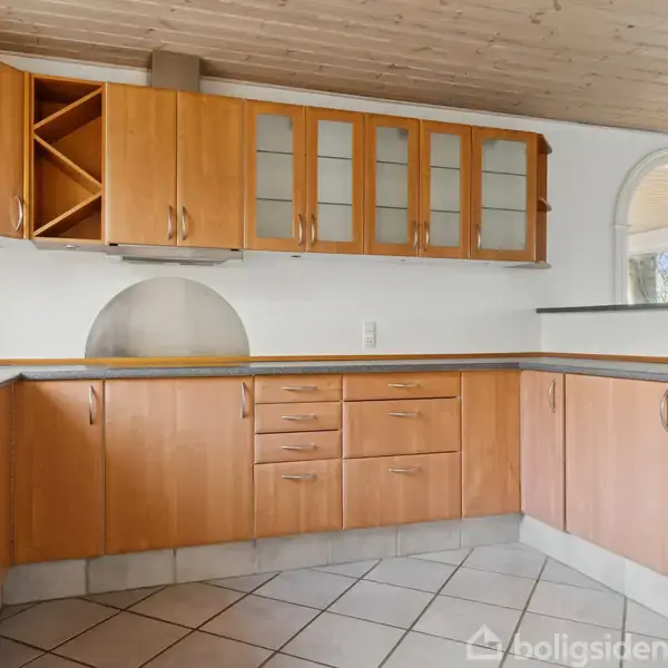 Køkken med træskabsfronter, glaslåger og indbygget vinkurv. Skabene hænger over en grå bordplade med firkantede, lysegrå gulvfliser. Buede vinduer i baggrunden lader lys strømme ind.