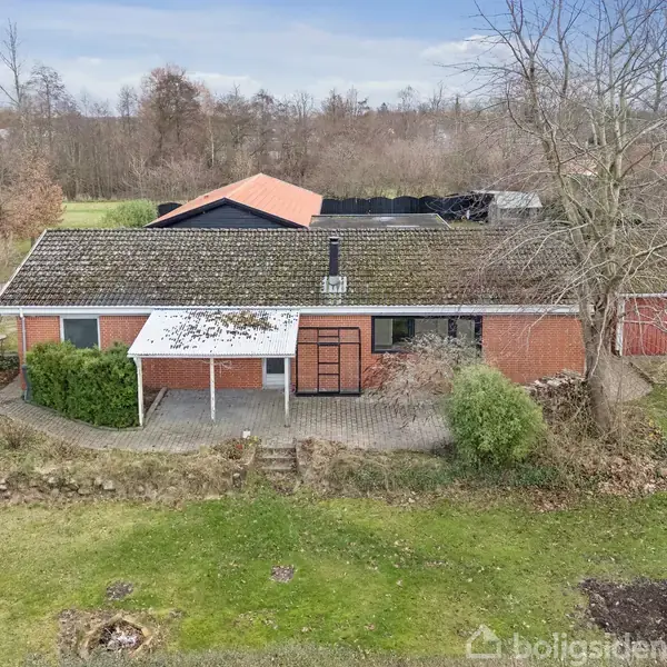 En rød murstensbygning med et dækket indgangsparti står midt i en have med træer og buske. Taget er skråt og området er omgivet af bevoksning og græsplæne.